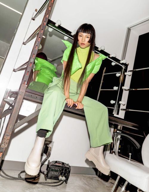 Young woman in bright green outfit seated on a metal ladder in a photo studio setting with lights and equipment visible.