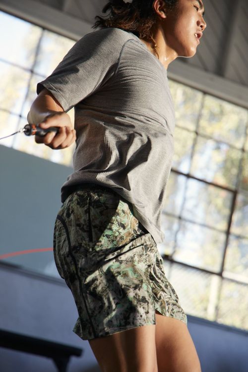 Athletic woman in a grey t-shirt and patterned shorts exercising with a jump rope in a bright indoor gym setting near windows.