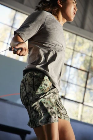 Athletic woman in a grey t-shirt and patterned shorts exercising with a jump rope in a bright indoor gym setting near windows.