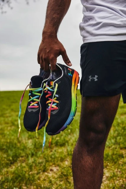 Close-up of a dark-skinned person holding black running shoes with rainbow laces, wearing black shorts, outdoors in natural light.