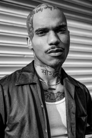 Striking portrait of a bald adult male with tattoos, wearing an open button-down shirt and silver chain, in a black and white composition.
