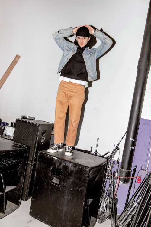 Man in denim jacket and black beanie standing on a speaker in an indoor setting with audio equipment and a white wall background.