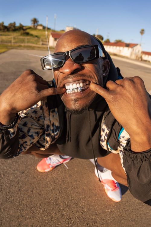 Stylish African-American man with grillz smiles outdoors, wearing sunglasses and an animal-print jacket in a low-angle fisheye lens shot.