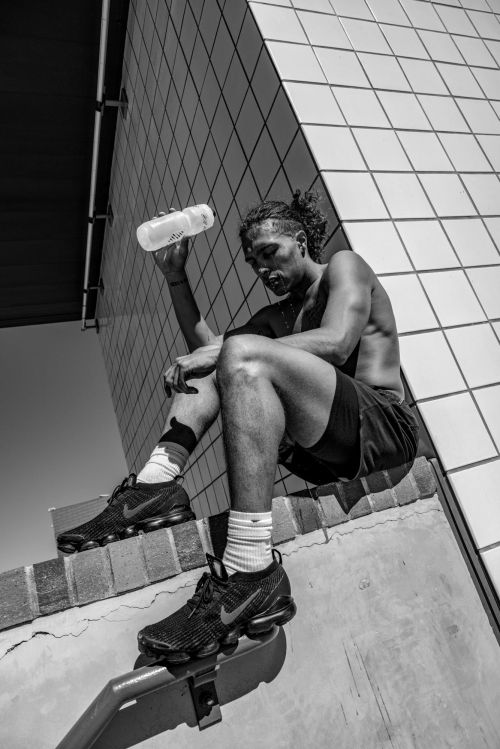 Black man drinking water outdoors while seated on a tiled ledge, wearing shorts and sneakers