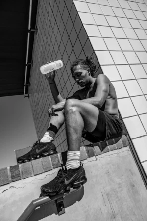Black man drinking water outdoors while seated on a tiled ledge, wearing shorts and sneakers