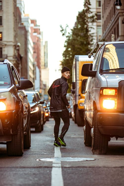 Adult male jogging through city street traffic wearing black beanie, jacket, and running shoes, medium shot, eye-level angle. Urban fitness shot.