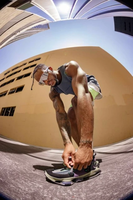 An athletic man ties his shoe on a city street under a bright sky. A fisheye lens captures nearby buildings and the surrounding urban environment.