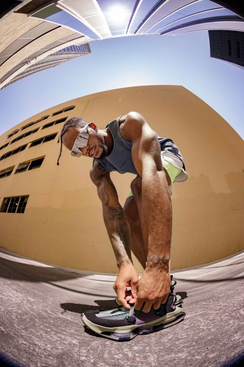 Eye-level shot of a man tying his shoe outdoors in daylight. The fisheye perspective captures a tall building and a clear sky overhead.