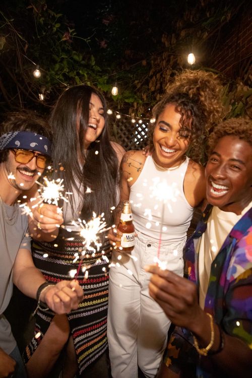 A group of four young adults smiles and laughs while holding sparklers outside at night, with a string of lights in the background.