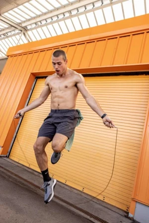 Shirtless man jumping rope outdoors in front of an orange garage-style door wearing athletic shorts, gray sneakers, and a fitness tracker.