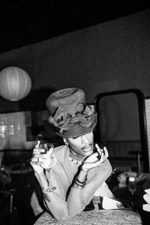 Black and white portrait of a person wearing an elaborate bow hat holding a wine glass in a dim interior by Andre L. Perry.