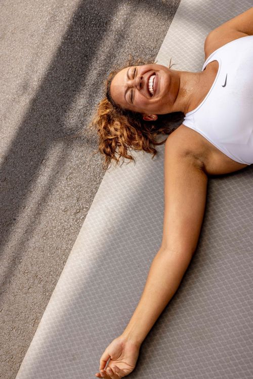 Athletic woman lying on a grey exercise mat outdoors, laughing, wearing a white tank top, after a workout, with sunlight and shadows.