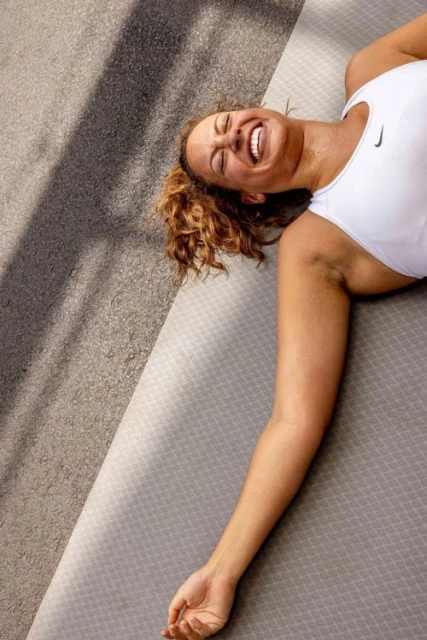 Athletic woman lying on a grey exercise mat outdoors, laughing, wearing a white tank top, after a workout, with sunlight and shadows.