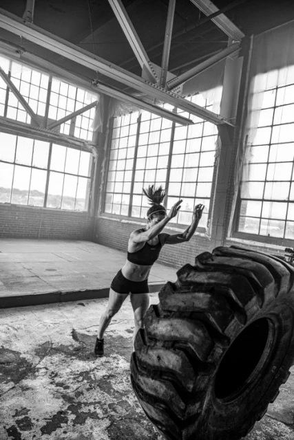 A woman in workout clothes flips a large tire in an industrial space, captured in black and white with natural light streaming through windows.