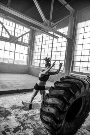 A woman in workout clothes flips a large tire in an industrial space, captured in black and white with natural light streaming through windows.