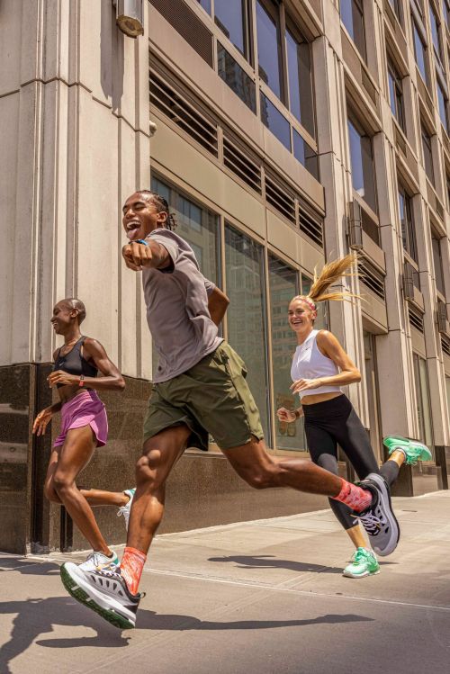 Three athletic adults run on a city sidewalk on a sunny day, showcasing fitness and urban outdoor activity with modern architecture in the background.