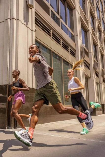 Three athletic adults run on a city sidewalk on a sunny day, showcasing fitness and urban outdoor activity with modern architecture in the background.