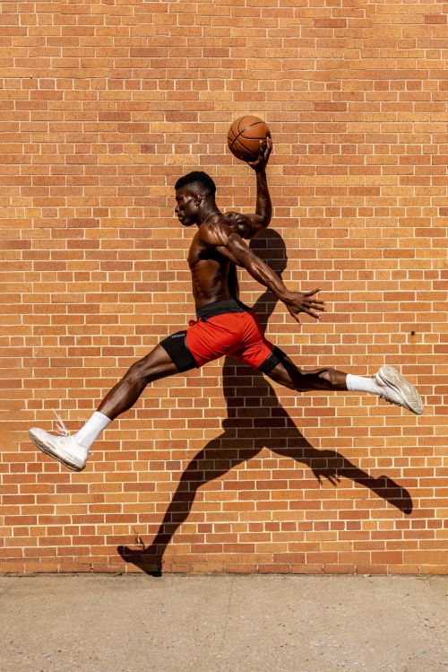 Athletic Black male in red shorts leaps dynamically with a basketball against a brick wall backdrop, casting a long shadow.
