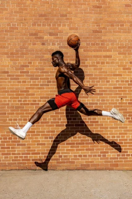 Athletic Black male in red shorts leaps dynamically with a basketball against a brick wall backdrop, casting a long shadow.