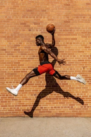 Athletic Black male in red shorts leaps dynamically with a basketball against a brick wall backdrop, casting a long shadow.