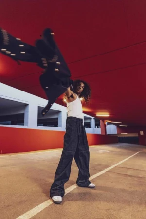 A person with long curly hair wearing a white tank top and wide-leg trousers takes off a dark jacket in a concrete parking garage.