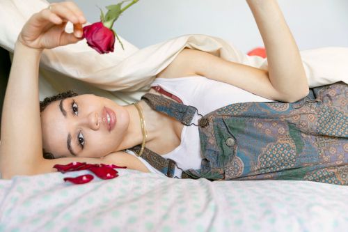 Woman lying on a bed holding a red rose near her face, wearing patterned overalls and a white tank top.
