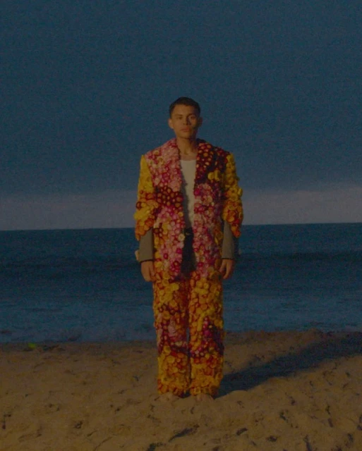 A full-length portrait showcases a slender, fair-skinned person on a beach, wearing a vibrant, floral-embellished suit under a twilight sky.