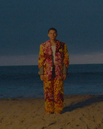 A full-length portrait showcases a slender, fair-skinned person on a beach, wearing a vibrant, floral-embellished suit under a twilight sky.