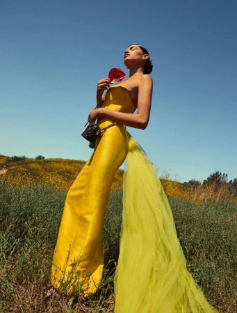 Model in a bright yellow jumpsuit holding a red flower outdoors against a clear blue sky, photographed by Brittany Bravo.
