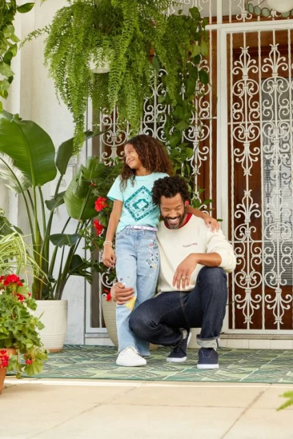 Father and daughter pose on a porch with plants. The child wears a graphic tee and jeans. The adult wears a cream sweatshirt and jeans.