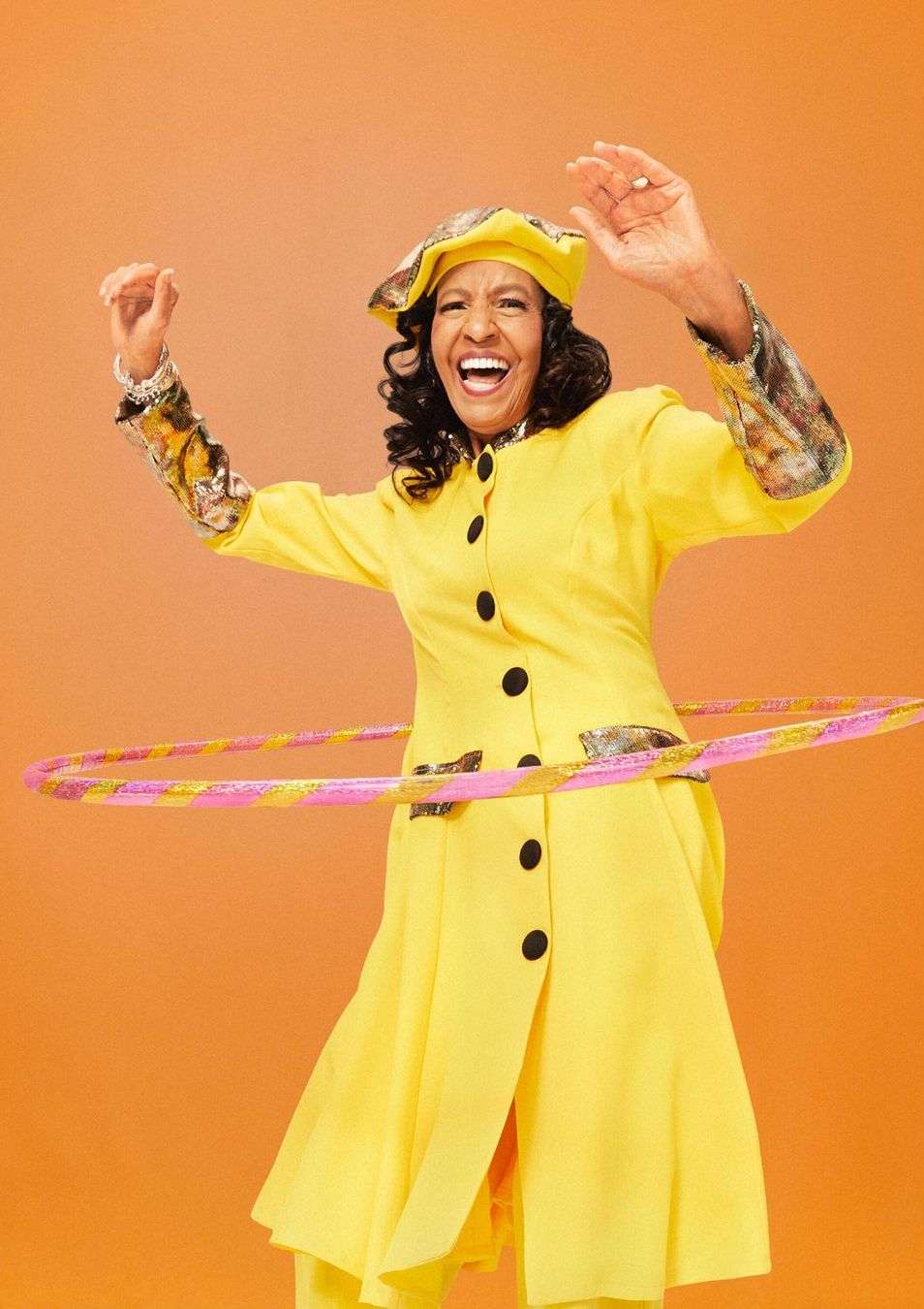 Cheerful woman with curly hair in yellow jacket and beret is joyfully hula-hooping against a plain orange backdrop in a studio shot.
