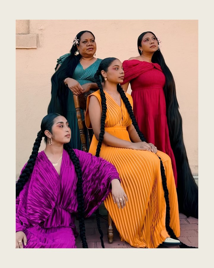 Group portrait of four women with long braided hair wearing colorful pleated gowns standing against a peach wall in the daylight.