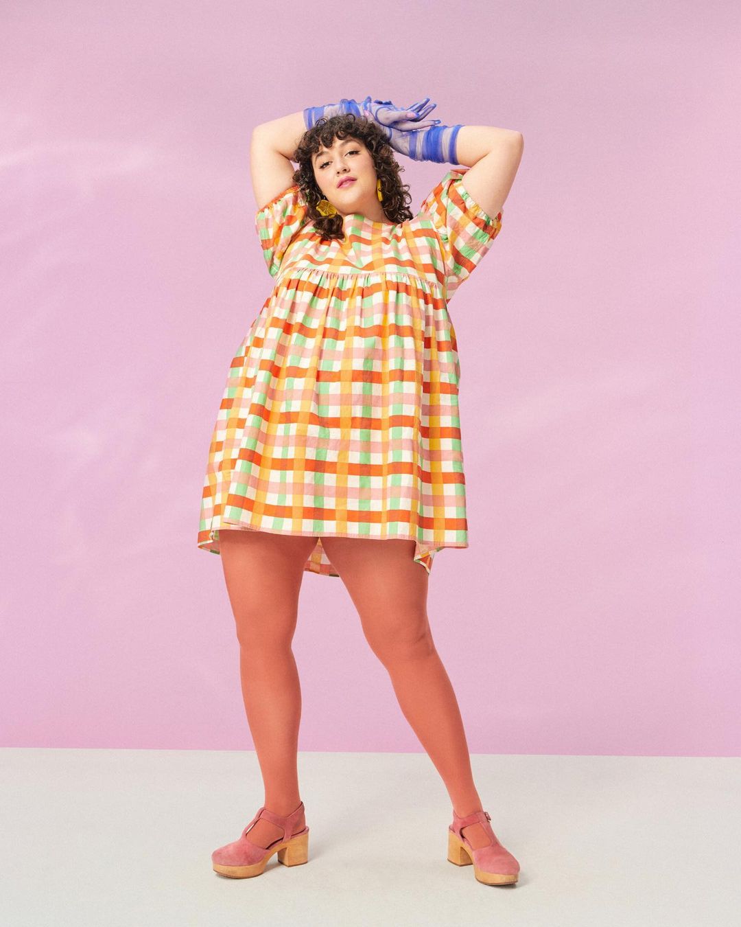 Woman in a colorful, plaid dress stands in a photo studio. She wears matching shoes and tights with striped gloves.