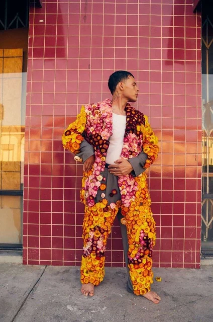 Man in floral suit posing against a red tiled wall outdoors. He is standing barefoot on a sidewalk, looking to his right.