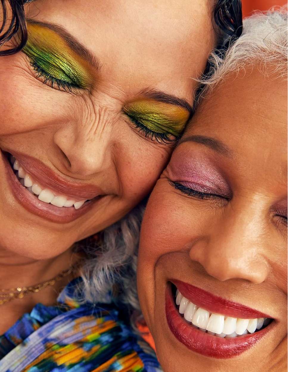 Close-up of two women with closed eyes, smiling, and wearing colourful eyeshadow, one in shades of green and the other in pink.