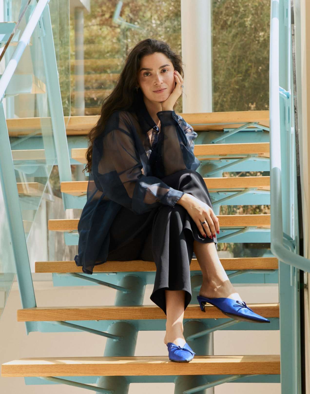 Brunette woman in sheer top and black pants sitting on light wood stairs indoors near a window with leafy trees outside.