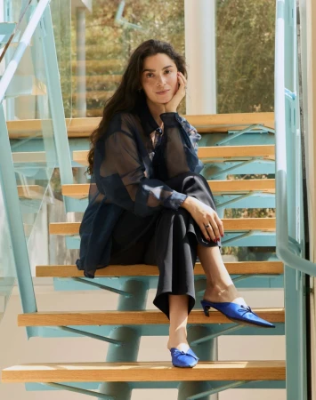 Brunette woman in sheer top and black pants sitting on light wood stairs indoors near a window with leafy trees outside.