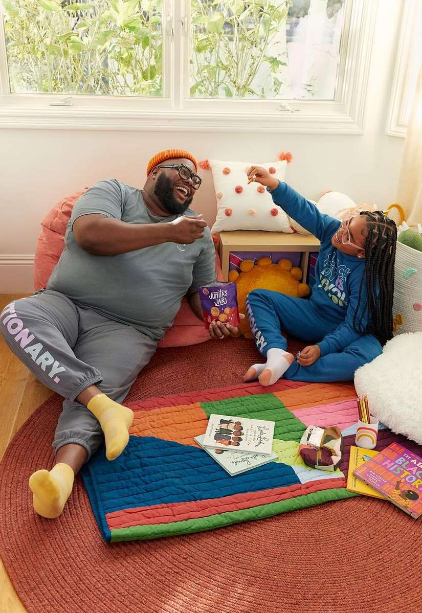 Cheerful father and daughter share snacks on a colorful rug with books and pillows in a cozy indoor setting by a window.