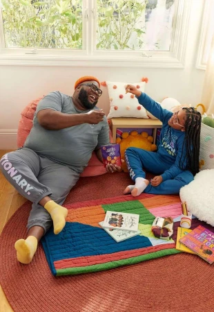 Cheerful father and daughter share snacks on a colorful rug with books and pillows in a cozy indoor setting by a window.