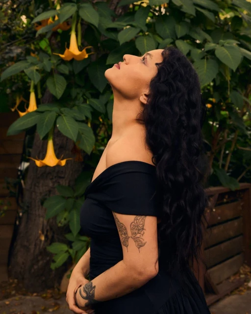 Adult woman in off-shoulder black dress gazes up at yellow Angel's Trumpet flowers amid lush green foliage in a garden setting.