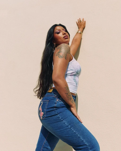 Portrait of a woman with long dark hair wearing jeans and a top against a light-yellow wall, with her arm raised and tattooed.