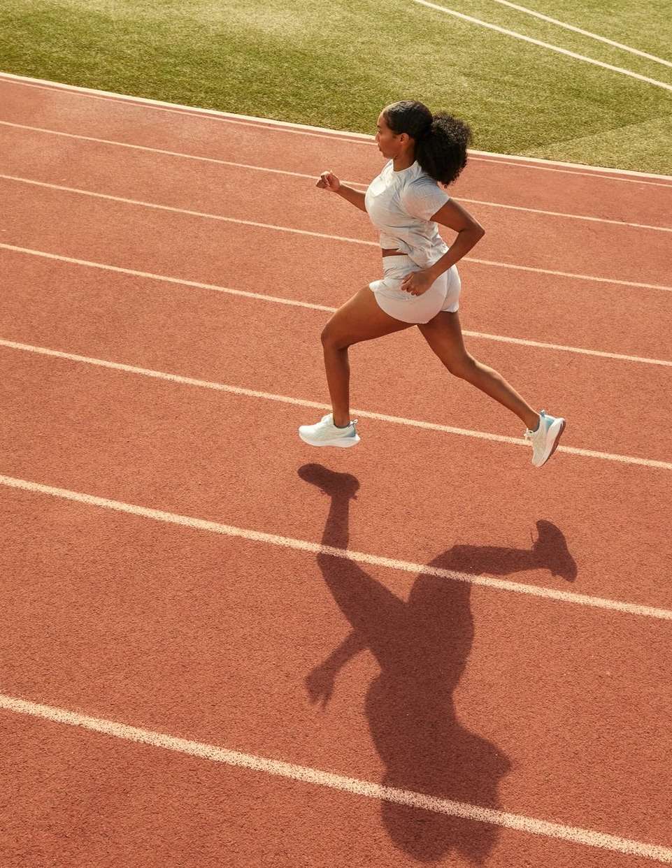 Overhead shot of an adult woman with dark hair running on a red running track in athletic wear on a sunny day outdoors.