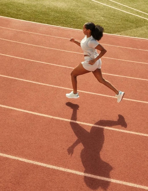 Overhead shot of an adult woman with dark hair running on a red running track in athletic wear on a sunny day outdoors.