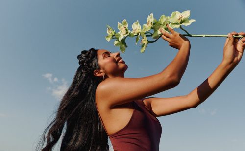 A woman with dark hair smiles and holds an orchid flower up to her face against a clear blue sky in an outdoor setting.