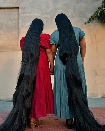 Two women with extremely long black hair standing outdoors, wearing red and teal dresses and holding hands.