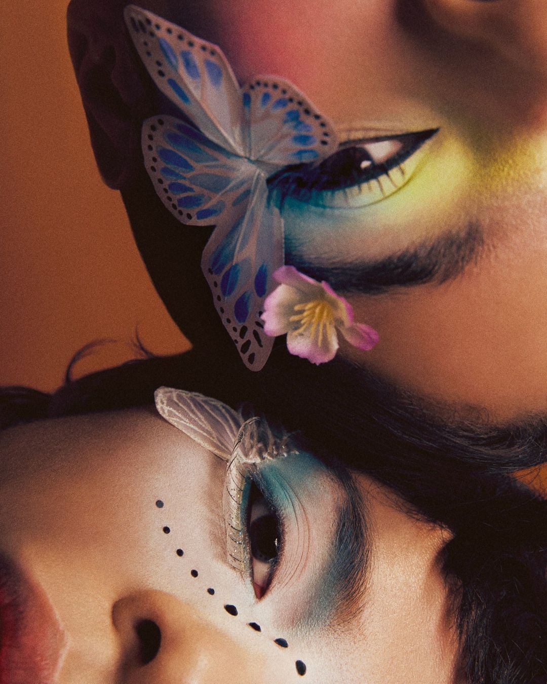 Aesthetic close-up studio portrait of two adult females with elaborate makeup, butterfly and floral embellishments, and dark hair against an orange backdrop.
