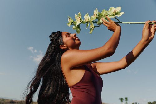 Woman in a swimsuit smiles and holds a stem of green and white orchids in an outdoor setting with a clear blue sky. 