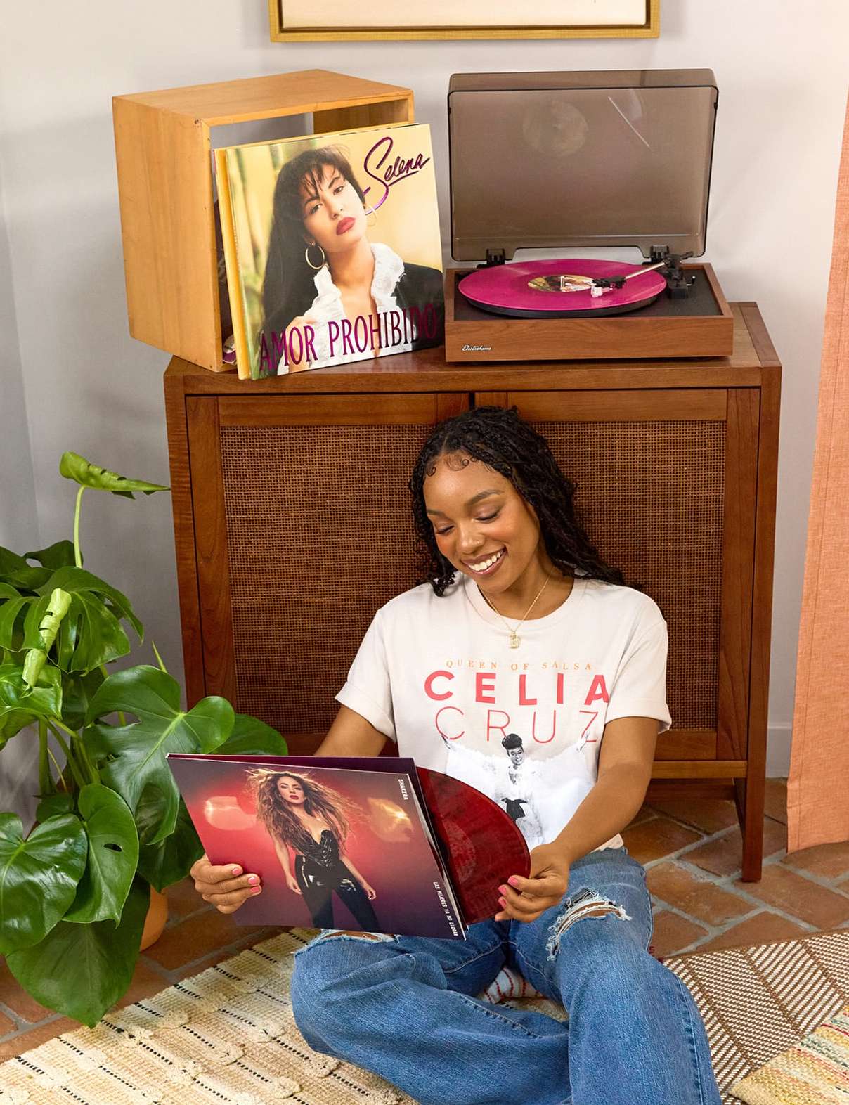 Woman sitting on a rug in a living room, holding a vinyl record in its cover, with a record player and a Selena record displayed on a console.