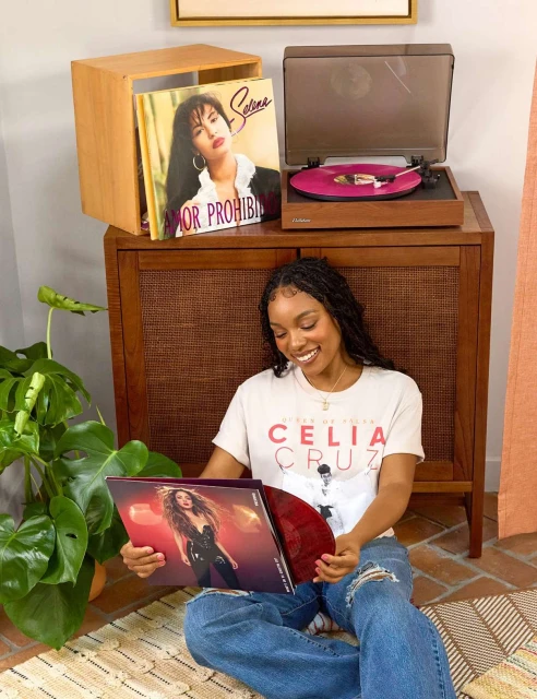 Woman sitting on a rug in a living room, holding a vinyl record in its cover, with a record player and a Selena record displayed on a console.