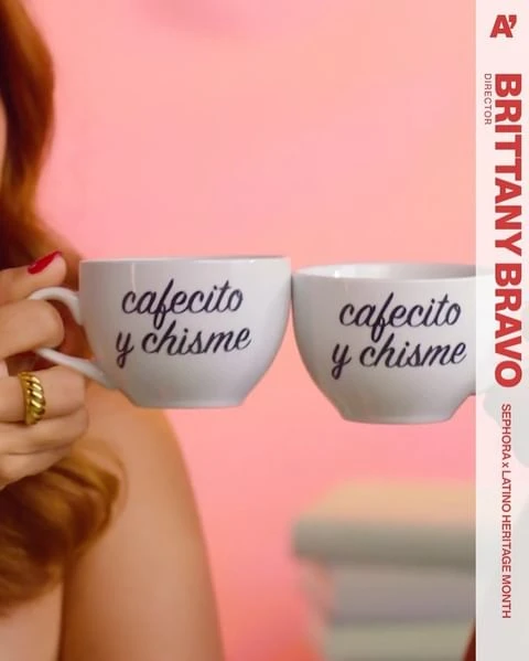 Two white ceramic mugs with the words 'cafecito y chisme' are held up against a pink background, partially obscuring a woman's face.